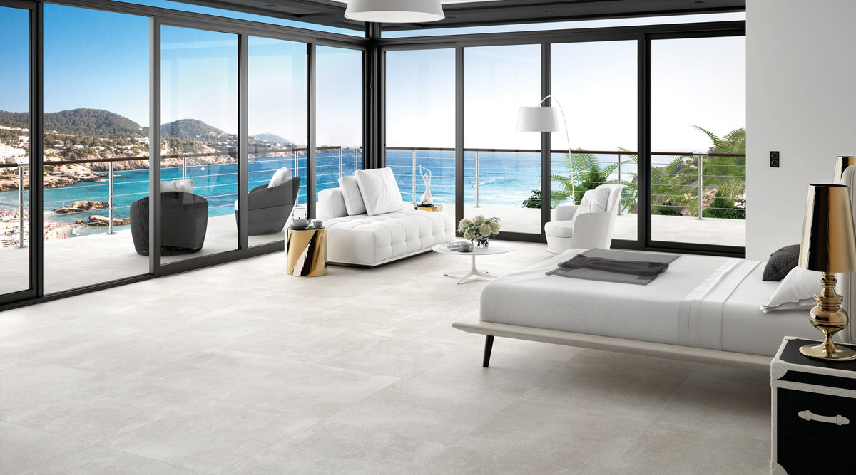 KTL Claire Gris 75x75 cm en dormitorio con cama y ventanal con vistas al mar, suelo porcelánico efecto cemento